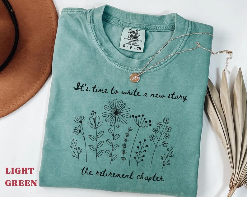 May include: A light green short-sleeved t-shirt featuring a black line art graphic of various wildflowers. The text reads, "It's time to write a new story" above the flowers and "the retirement chapter" below the flowers in a cursive font.