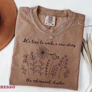 May include: An espresso-colored short-sleeve t-shirt with a crew neckline. The shirt features a black line-art graphic of various wildflowers and the text: It's time to write a new story, the retirement chapter. A silver-toned necklace rests on the collar.