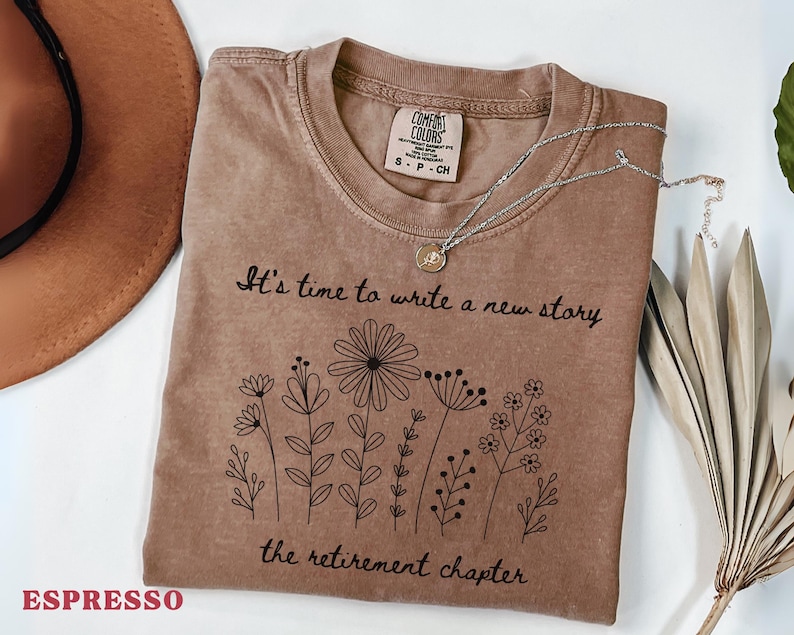 May include: An espresso-colored short-sleeve t-shirt with a crew neckline. The shirt features a black line-art graphic of various wildflowers and the text: It's time to write a new story, the retirement chapter. A silver-toned necklace rests on the collar.