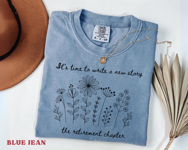 May include: A blue jean colored t-shirt with a black graphic print of various wildflowers and the text It's time to write a new story the retirement chapter. The shirt features a Comfort Colors brand label at the inner neckline and a gold necklace.