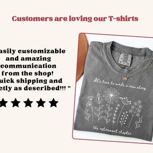 May include: A charcoal gray t-shirt featuring a white line art graphic of various wildflowers. The text on the shirt reads, "It's time to write a new story" above the flowers and "the retirement chapter" below them. The shirt has a crew neckline.