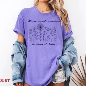 May include: A violet short-sleeved t-shirt featuring a black line-art illustration of various wildflowers. The text "It's time to write a new story" is printed above the flowers, and "the retirement chapter" is printed below the floral design.