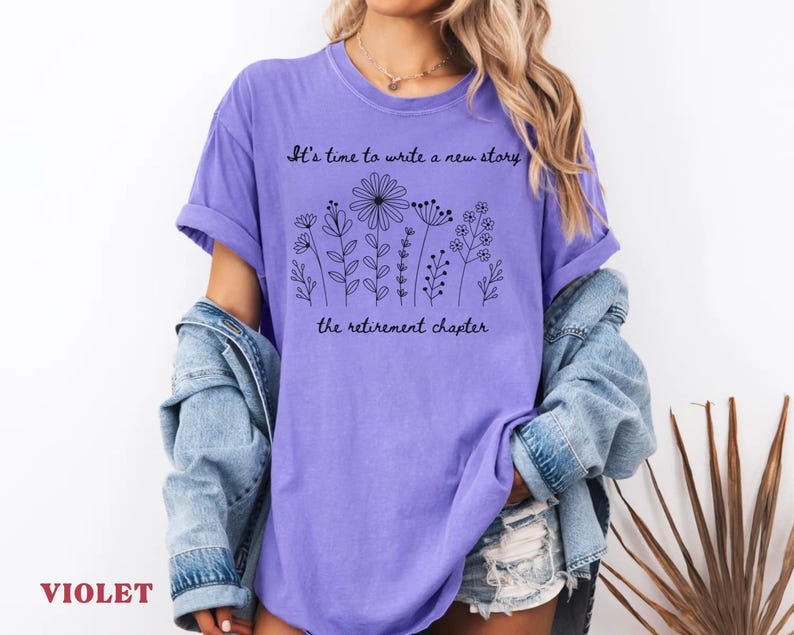 May include: A violet short-sleeved t-shirt featuring a black line-art illustration of various wildflowers. The text "It's time to write a new story" is printed above the flowers, and "the retirement chapter" is printed below the floral design.