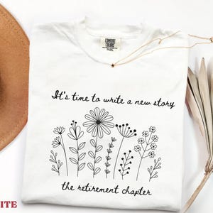 May include: A white t-shirt featuring a black line art illustration of various wildflowers. The text "It's time to write a new story" is printed above the flowers, and "the retirement chapter" is printed below the floral design in black.