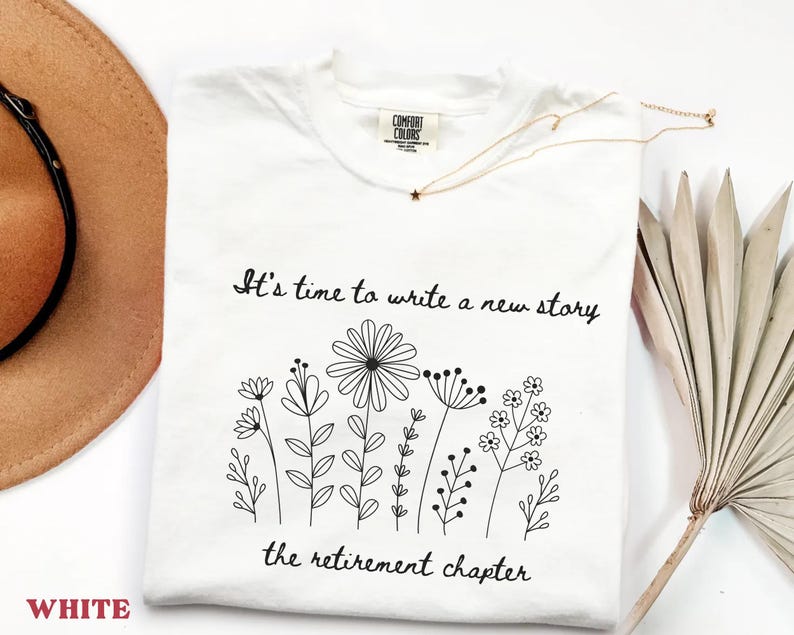 May include: A white t-shirt featuring a black line art illustration of various wildflowers. The text "It's time to write a new story" is printed above the flowers, and "the retirement chapter" is printed below the floral design in black.