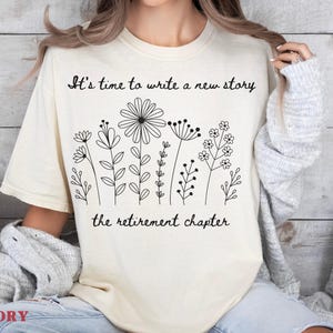 May include: An ivory t-shirt featuring a black line art graphic of various wildflowers. The text above the flowers reads, "It's time to write a new story," and the text below the flowers reads, "the retirement chapter." The shirt has short sleeves.