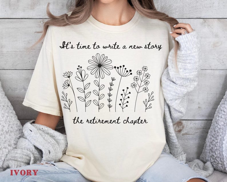 May include: An ivory t-shirt featuring a black line art graphic of various wildflowers. The text above the flowers reads, "It's time to write a new story," and the text below the flowers reads, "the retirement chapter." The shirt has short sleeves.