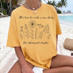 May include: A mustard yellow short-sleeve t-shirt featuring a black line art illustration of various wildflowers. The text "It's time to write a new story" is printed above the flowers, and "the retirement chapter" is printed below them.