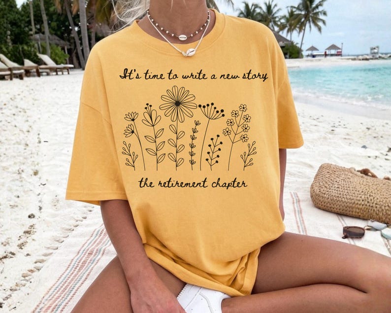 May include: A mustard yellow short-sleeve t-shirt featuring a black line art illustration of various wildflowers. The text "It's time to write a new story" is printed above the flowers, and "the retirement chapter" is printed below them.
