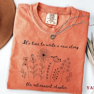 May include: A coral-colored short-sleeve t-shirt with a crew neckline. The shirt features a black line-art graphic of various wildflowers and the printed text: It's time to write a new story, the retirement chapter. A Comfort Colors brand label is visible.