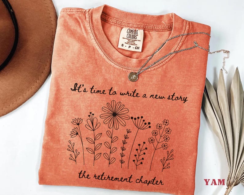 May include: A coral-colored short-sleeve t-shirt with a crew neckline. The shirt features a black line-art graphic of various wildflowers and the printed text: It's time to write a new story, the retirement chapter. A Comfort Colors brand label is visible.