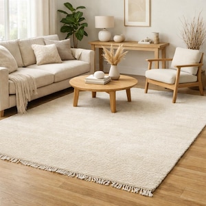 May include: A neutral-toned living room rug with a fringed edge. The rug is placed on a hardwood floor, with a sofa, armchair, and coffee table visible in the background. The room is decorated in a minimalist style.