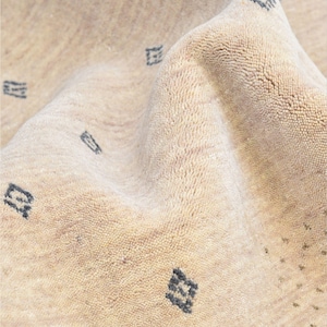 Puede incluir: Primer plano de una alfombra beige con una superficie texturizada y un patr&oacute;n sutil. La alfombra presenta peque&ntilde;as formas geom&eacute;tricas de color gris oscuro dispersas por su superficie. La tela est&aacute; doblada, mostrando la profundidad y la textura del material.