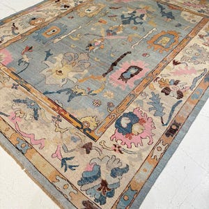Könnte beinhalten: Ein großer, rechteckiger Teppich mit einem hellblauen Mittelfeld und einem detaillierten Rand. Der Teppich weist florale und geometrische Muster in Rosa-, Orange-, Beige- und Blautönen auf. Das Design des Teppichs ist aufwendig und die Farben sind weich.