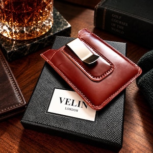 Può includere: Un portafoglio fermasoldi in pelle rosso scuro con una clip in metallo argentato lucido, appoggiato su una scatola regalo nera testurizzata con la scritta VELIN LONDON in lettere maiuscole nere, con cuciture visibili lungo i bordi della pelle.