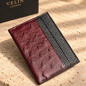 May include: A rectangular card holder featuring a burgundy ostrich-textured leather panel on the left and a black pebbled leather panel on the right with vertical card slots. The black box in the background displays the gold text VELIN LONDON.