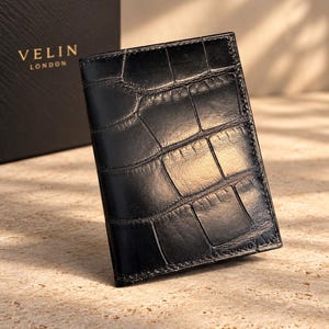 May include: A black card holder with a glossy crocodile skin texture. The rectangular accessory features a vertical orientation and visible stitching along the edges. A black box with the gold text VELIN LONDON is positioned behind the card holder.
