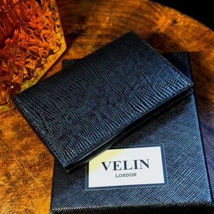 May include: A black textured leather wallet and its matching box. The wallet is open, showing the inside. The box features a white label with the text "VELIN LONDON". The background is a dark wooden surface.