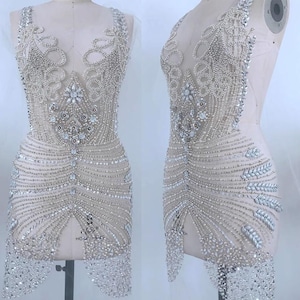 May include: A sleeveless mini dress featuring a sheer nude base adorned with intricate silver beadwork, clear crystals, and sequins arranged in swirling patterns and geometric lines, with a deep neckline and a heavily embellished fringe hemline.