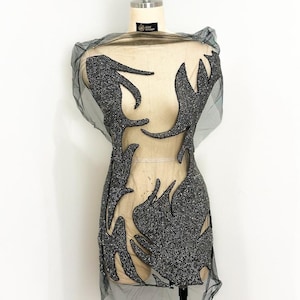 May include: A sheer black tulle garment featuring intricate, flame-like appliqué patterns densely covered in shimmering silver beads. The design creates a bold, high-contrast visual effect against the translucent fabric, showcasing detailed craftsmanship.