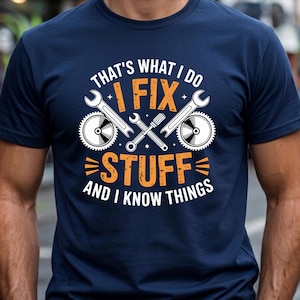 May include: A navy blue short-sleeve t-shirt featuring a graphic print with the text "THAT'S WHAT I DO I FIX STUFF AND I KNOW THINGS" in white and orange. The design includes illustrations of two wrenches, two circular saw blades, and a screwdriver.