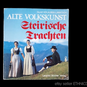 May include: A book cover with the title "Alte Volkskunst Steirische Trachten" in red and black lettering. The cover features a mountain landscape with three people wearing traditional Austrian clothing.