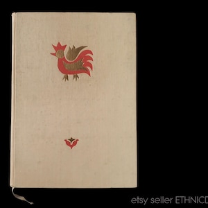 May include: A vintage book with a cream-colored cover featuring a red and gold rooster illustration at the top and a red floral design at the bottom. The book has a ribbon bookmark and is set against a black background.