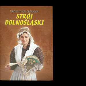 May include: A book cover with the title "Piękno stroju ludowego STRÓJ DOLNOŚLĄSKI" in yellow text on a brown background. A woman in a traditional Polish folk costume is holding a hat in her hands.