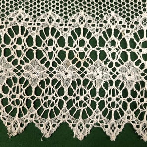 May include: Close-up of a cream-colored lace fabric with an intricate openwork design. The lace features a repeating pattern of geometric shapes and scalloped edges, creating a delicate and textured appearance. The background is a dark green.