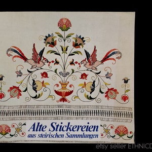 May include: A vintage textile with colourful embroidered floral and bird designs. The artwork features a central vase with flowers, flanked by birds with long tail feathers. The text "Alte Stickereien" is visible at the bottom, indicating the item's origin.
