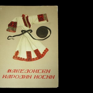 May include: A book cover with a white embroidered shirt with red and black trim, a black belt, and a black hat. The text on the cover reads "МАКЕДОНСКИ НАРОДНИ НОСИИ".