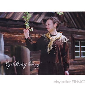 May include: A woman wearing a brown dress with a yellow scarf and a floral print on the dress. The woman is standing in front of a wooden building with a window. The text "Opoloski strój ludowy" is written in white on the bottom left corner of the image.