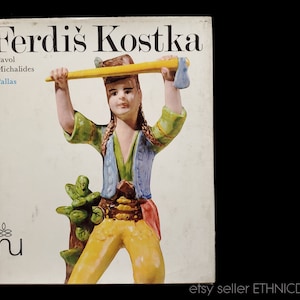 Könnte beinhalten: Ein Buchcover mit einer bunten Keramikfigur einer Person, die einen gelben Stock hält. Der Buchtitel lautet "Ferdiš Kostka" mit dem Namen des Autors "Pavol Michalides" und dem Verlag "Pallas" darunter. Das Buchcover ist weiß mit einem blauen und grünen Design.