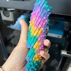 May include: A colorful, 3D-printed object with a textured, ridged surface. The object is a gradient of colors, including pink, yellow, green, and blue. The object is held in a hand, with a 3D printer visible in the background.