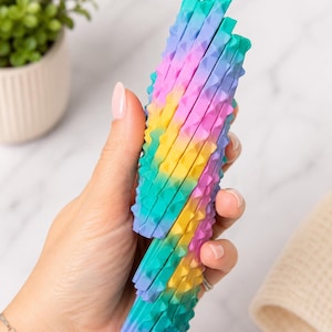 May include: A colorful, stackable sensory toy in shades of teal, yellow, pink, and purple. The toy has a textured, studded surface and is held in a hand. The background includes a small potted plant and a marble surface.
