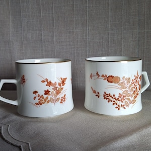 May include: Two white ceramic mugs with a floral design in shades of orange and brown. The mugs have a gold rim and a unique square handle. The mugs are placed on a neutral-colored surface.