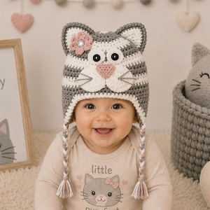May include: A baby wearing a gray and white crochet cat hat with a pink flower. The baby is wearing a light-colored onesie with a cat graphic and the words "little purrfect". A framed picture says "So purrfectly loved".