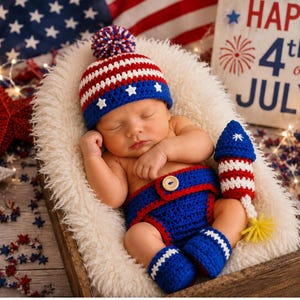 May include: A newborn baby wearing a patriotic crochet outfit, including a hat, diaper cover, and booties in red, white, and blue. The baby is nestled in a wooden basket with a fluffy white lining, with a decorative rocket and a sign that says "Happy 4th of July".