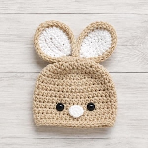 May include: A tan crochet beanie featuring two upright rabbit ears with white centers. The hat includes two black circular eyes and a small white circular nose stitched onto the front, creating a simple animal face design made of yarn.