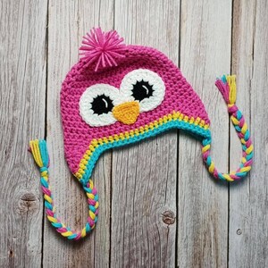 Puede incluir: Un gorro de b&uacute;ho rosa tejido a ganchillo con ojos blancos, pupilas negras y un pico naranja. El gorro tiene un pomp&oacute;n rosa en la parte superior y orejeras trenzadas con hebras amarillas, azules y rosas. El gorro es para ni&ntilde;os.