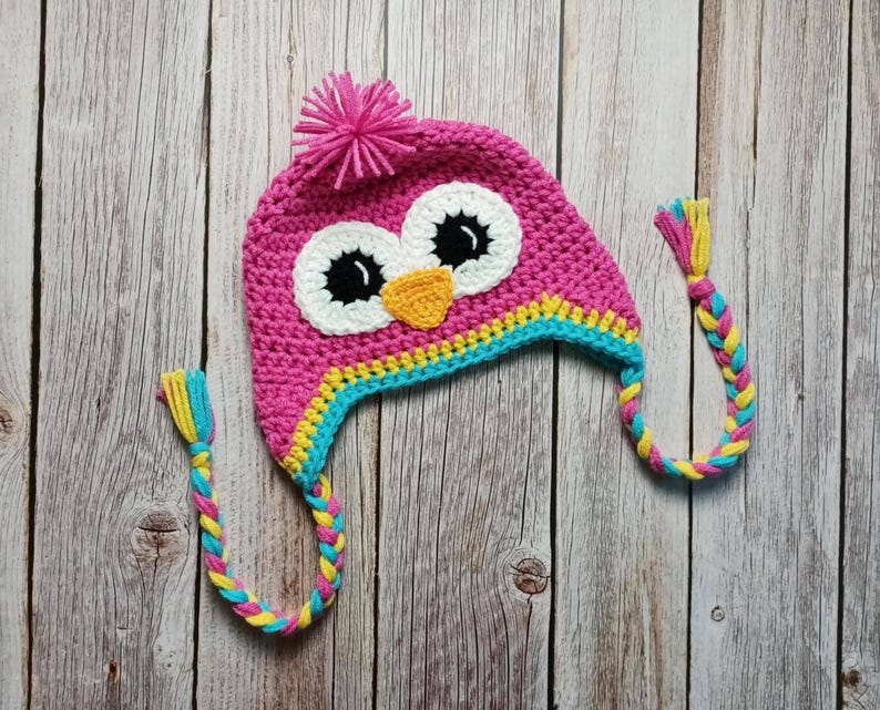 Puede incluir: Un gorro de b&uacute;ho rosa tejido a ganchillo con ojos blancos, pupilas negras y un pico naranja. El gorro tiene un pomp&oacute;n rosa en la parte superior y orejeras trenzadas con hebras amarillas, azules y rosas. El gorro es para ni&ntilde;os.