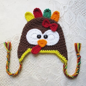 May include: A brown crocheted turkey hat with a red beak, white eyes, and a yellow brim. The hat has a long, braided cord in red, yellow, and green.