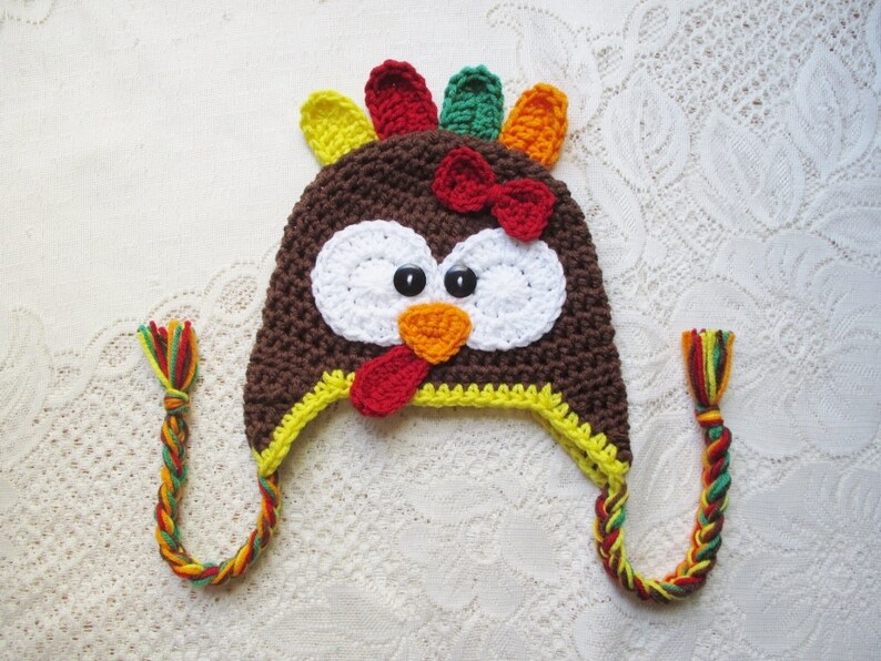 May include: A brown crocheted turkey hat with a red beak, white eyes, and a yellow brim. The hat has a long, braided cord in red, yellow, and green.