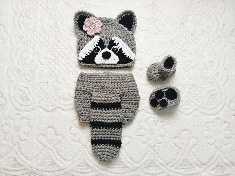 K&ouml;nnte beinhalten: Geh&auml;keltes Babykost&uuml;m-Set im Waschb&auml;r-Design in Grau und Schwarz. Das Set besteht aus einem Hut mit rosa Blume, einer Windel mit gestreiftem Schwanz und Babyschuhen mit Pfotenpolstern.