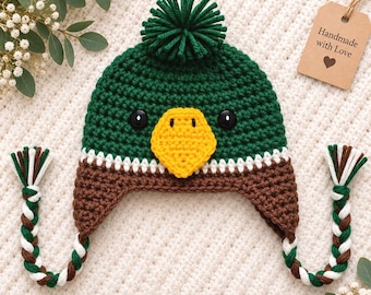 May include: A dark green crochet hat featuring a pom-pom on top, a bright yellow beak, and two black button eyes. The hat has brown ear flaps with braided tassels in green, brown, and white. A brown tag reads Handmade with Love.