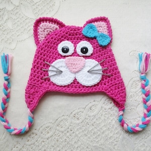 May include: A handmade, hot pink crochet cat hat with a light blue bow, white and pink accents, and braided ear ties. The hat features large black eyes, a pink nose, and white whiskers. A whimsical and cozy accessory.