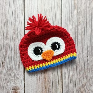 Puede incluir: Un gorro de b&uacute;ho rojo tejido a ganchillo con ojos blancos, pupilas negras y un pico naranja. El gorro tiene un pomp&oacute;n rojo en la parte superior y un ribete azul, amarillo y azul. El gorro est&aacute; dise&ntilde;ado para beb&eacute;s y ni&ntilde;os peque&ntilde;os.