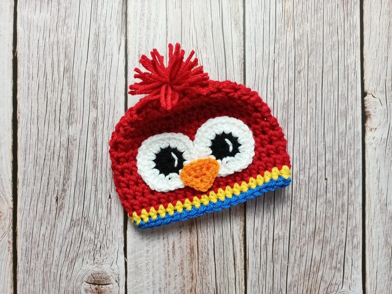 Puede incluir: Un gorro de b&uacute;ho rojo tejido a ganchillo con ojos blancos, pupilas negras y un pico naranja. El gorro tiene un pomp&oacute;n rojo en la parte superior y un ribete azul, amarillo y azul. El gorro est&aacute; dise&ntilde;ado para beb&eacute;s y ni&ntilde;os peque&ntilde;os.