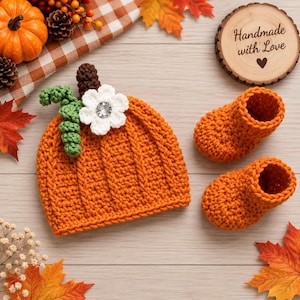 May include: A crochet pumpkin hat in orange with a brown stem, green curly vine, and a white flower with a central crystal accent, accompanied by a pair of matching orange crochet booties. A wooden sign reads Handmade with Love.
