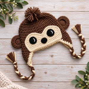 May include: A brown and cream crocheted hat shaped like a monkey face, featuring a dark brown yarn pom-pom on top, two round ears, and two long braided tassels made of alternating brown and cream yarn strands hanging from the sides.